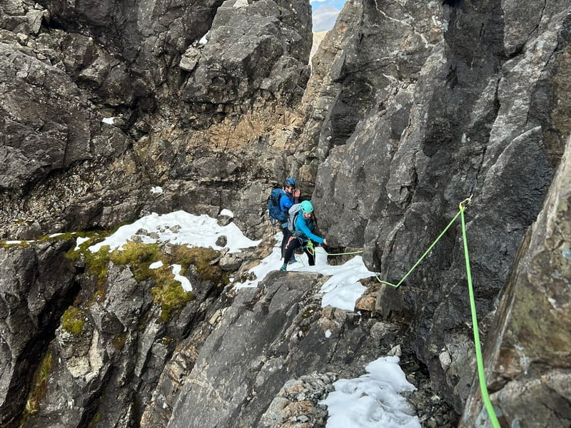2 people scrambling up a rocky ledge in snow, assisted by ropes, in the Lake District